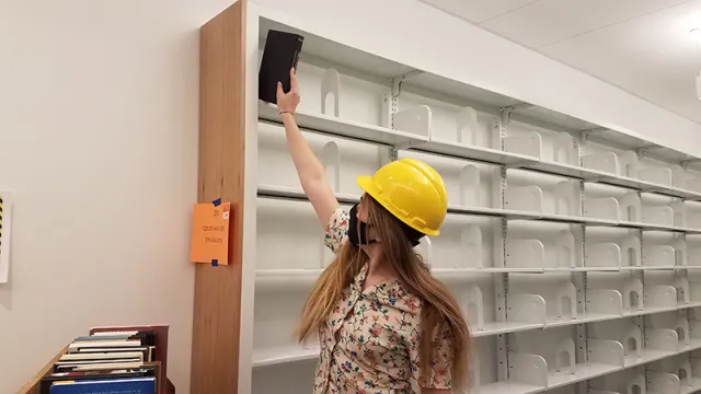 young woman with long blond hair putting a book high on a metal book shelf